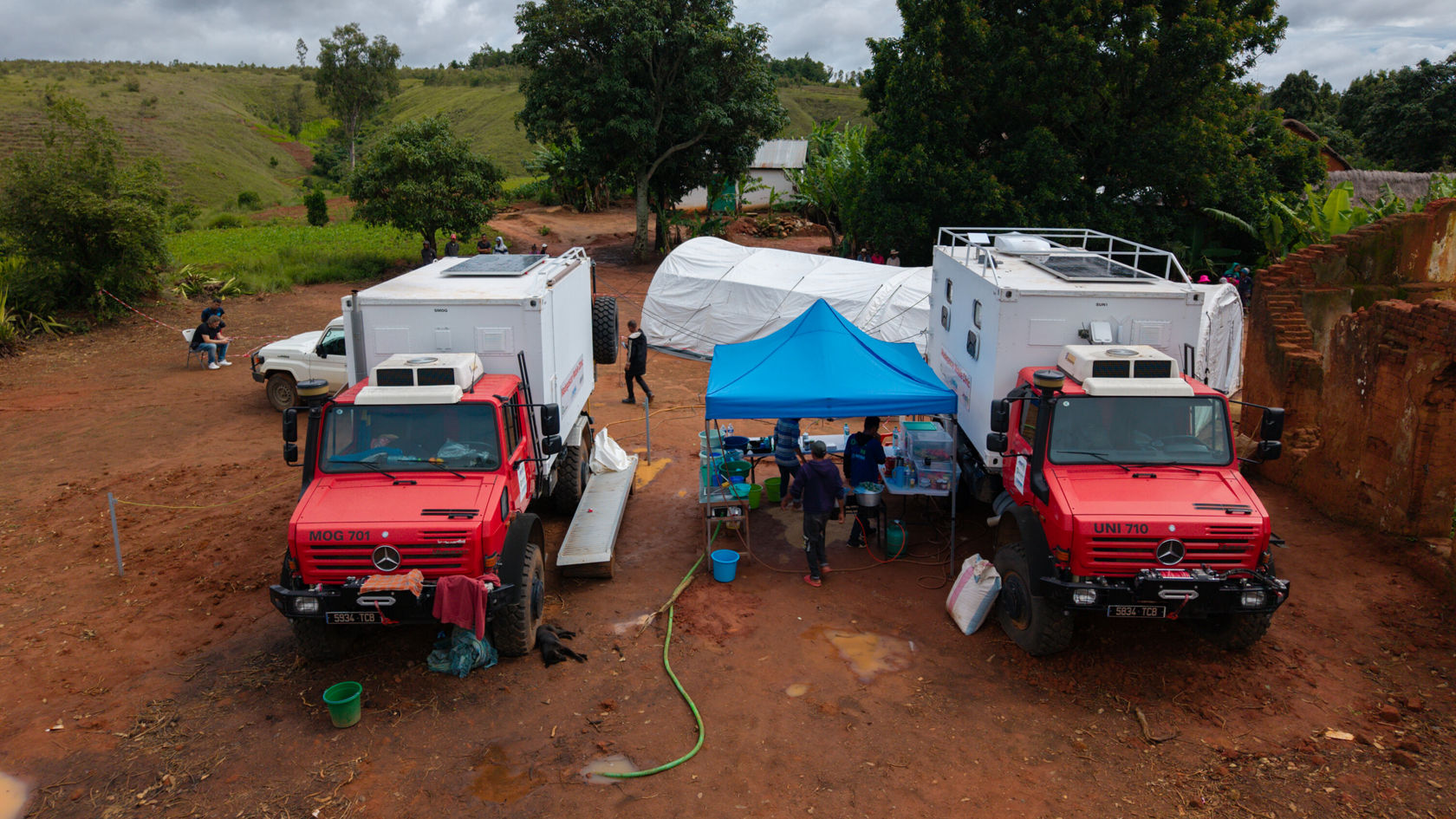 Deux Unimog Mercedes‑Benz rouges se trouvent à côté d'une tente bleue, dans laquelle des personnes travaillent avec du matériel. À l'arrière-plan, des collines vertes sont visibles.
