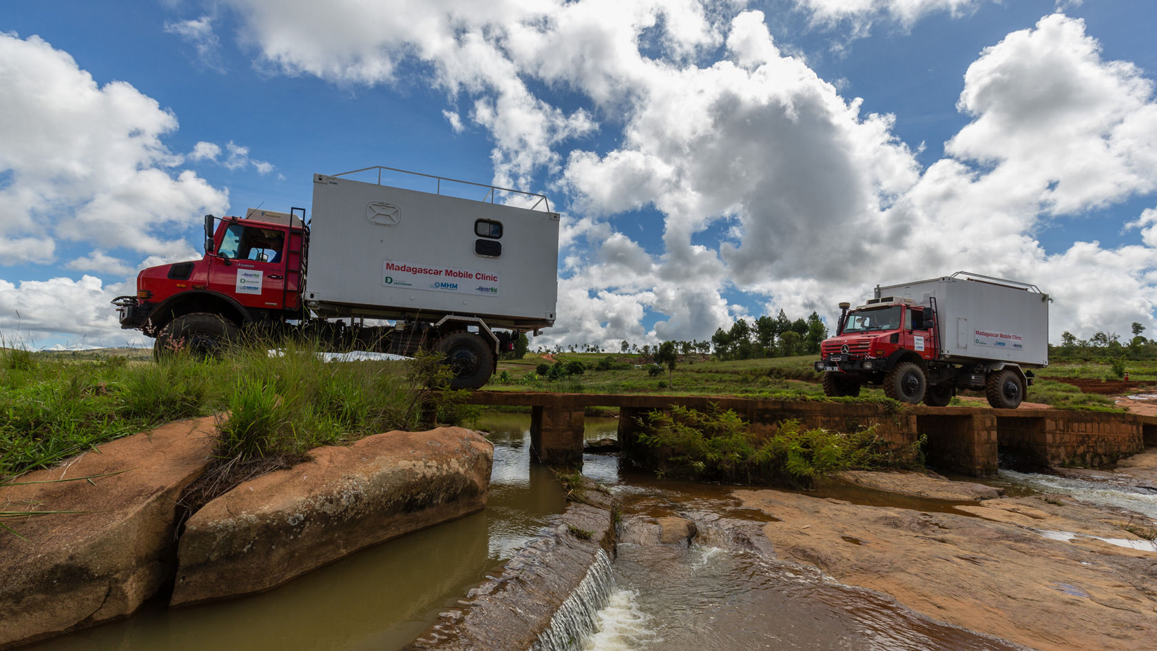 Deux véhicules Mercedes‑Benz, équipés pour le tout-terrain, traversent un pont en pierre dans une campagne, entourés de prairies verdoyantes et sous un ciel parsemé de nuages.