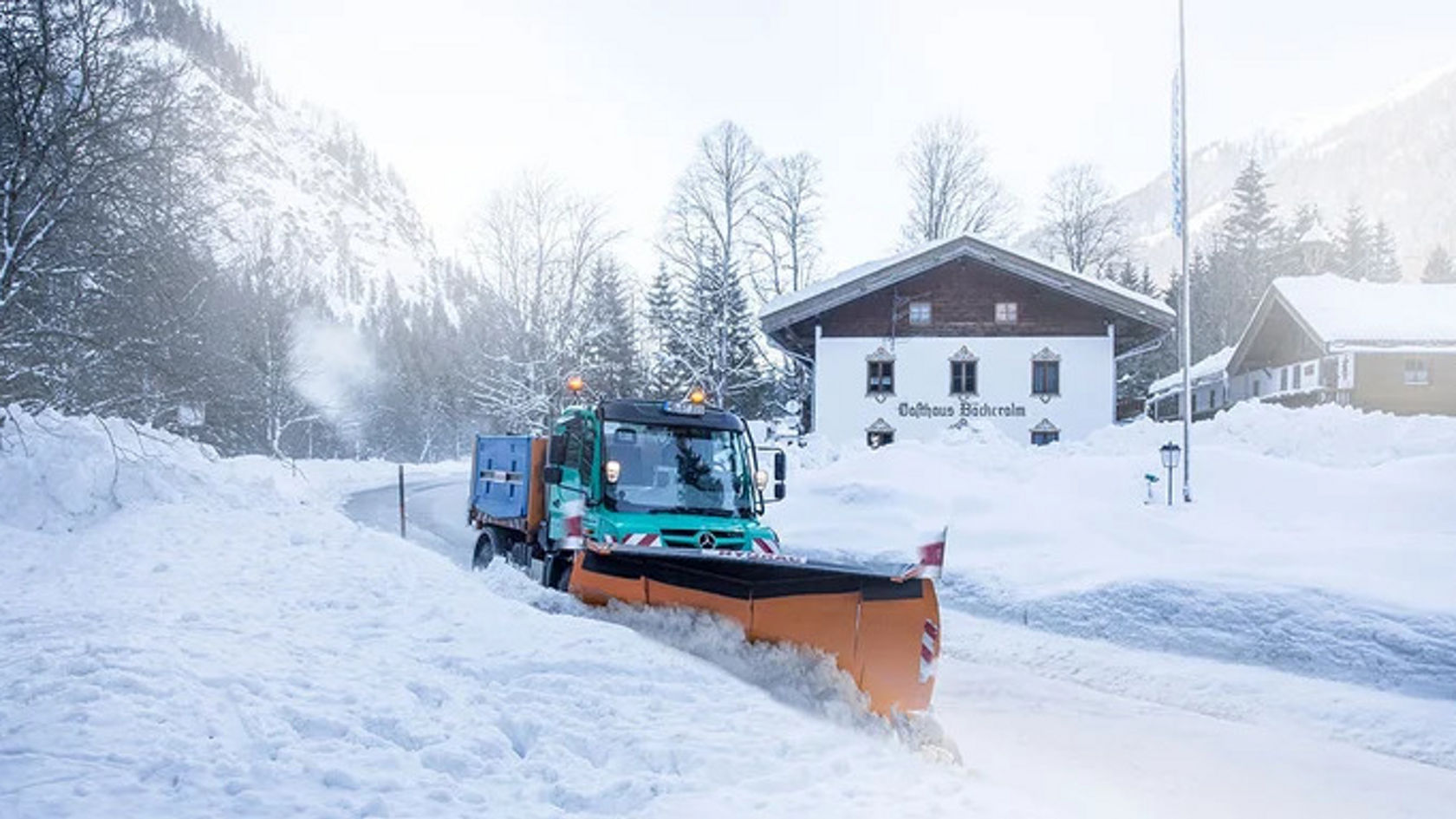 Un chasse-neige Mercedes-Benz dégage une route enneigée devant une maison alpine, entourée d'arbres recouverts de neige et de montagnes enneigées.