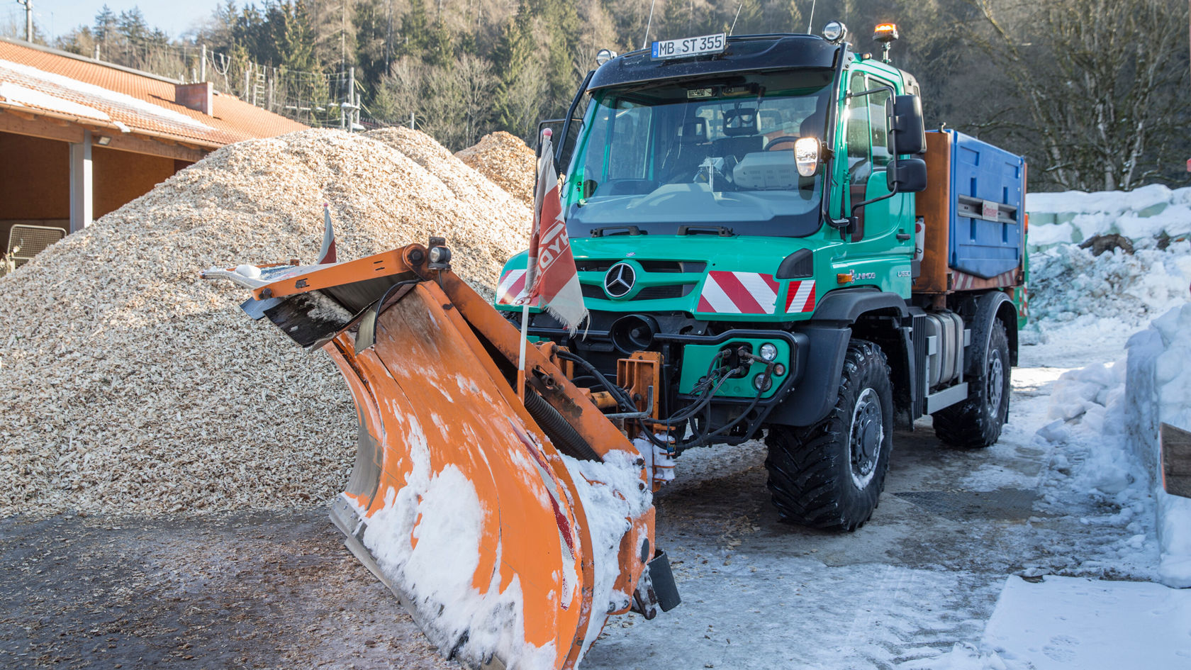 Un véhicule de transport Mercedes‑Benz équipé d'une lame biaise orange est stationné sur un parking recouvert de neige à côté d'un tas de bois.