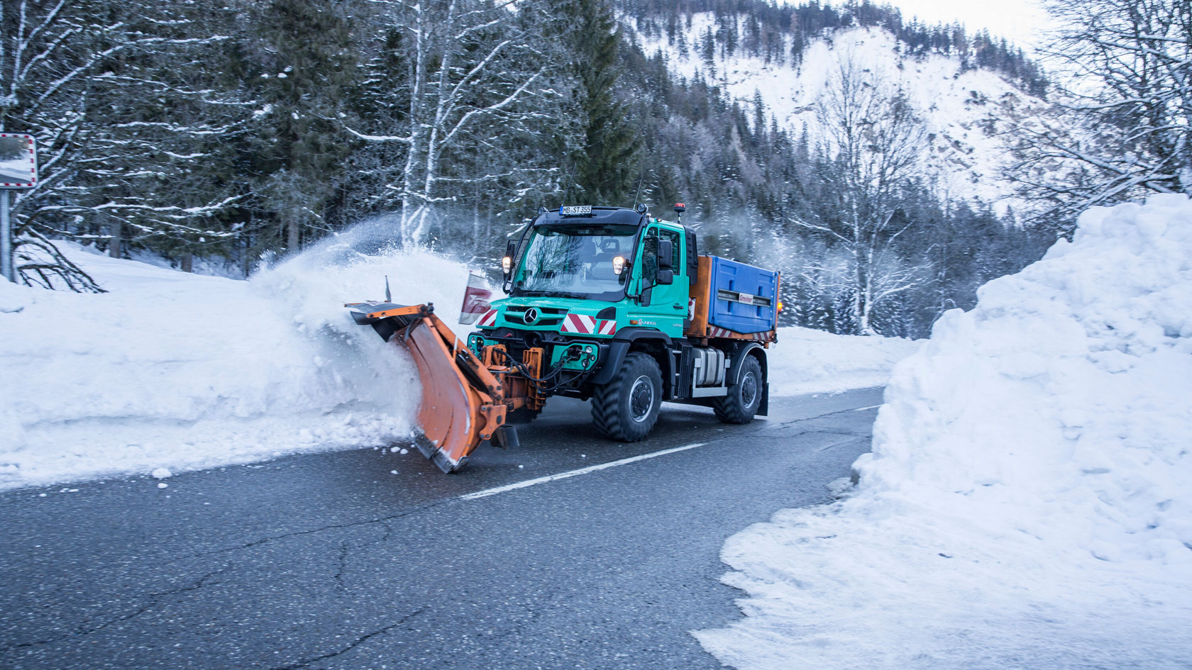 Un chasse-neige dégage une route enneigée dans la forêt, tandis que de hautes congères sont visibles de part et d'autre.