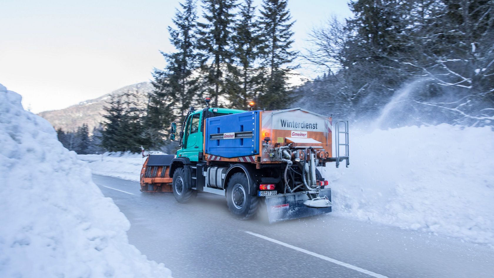 Un véhicule de service hivernal Mercedes‑Benz déblaye une route enneigée, tandis que la neige est repoussée sur le côté par la lame de déneigement. Des arbres et des montagnes sont visibles en arrière-plan.