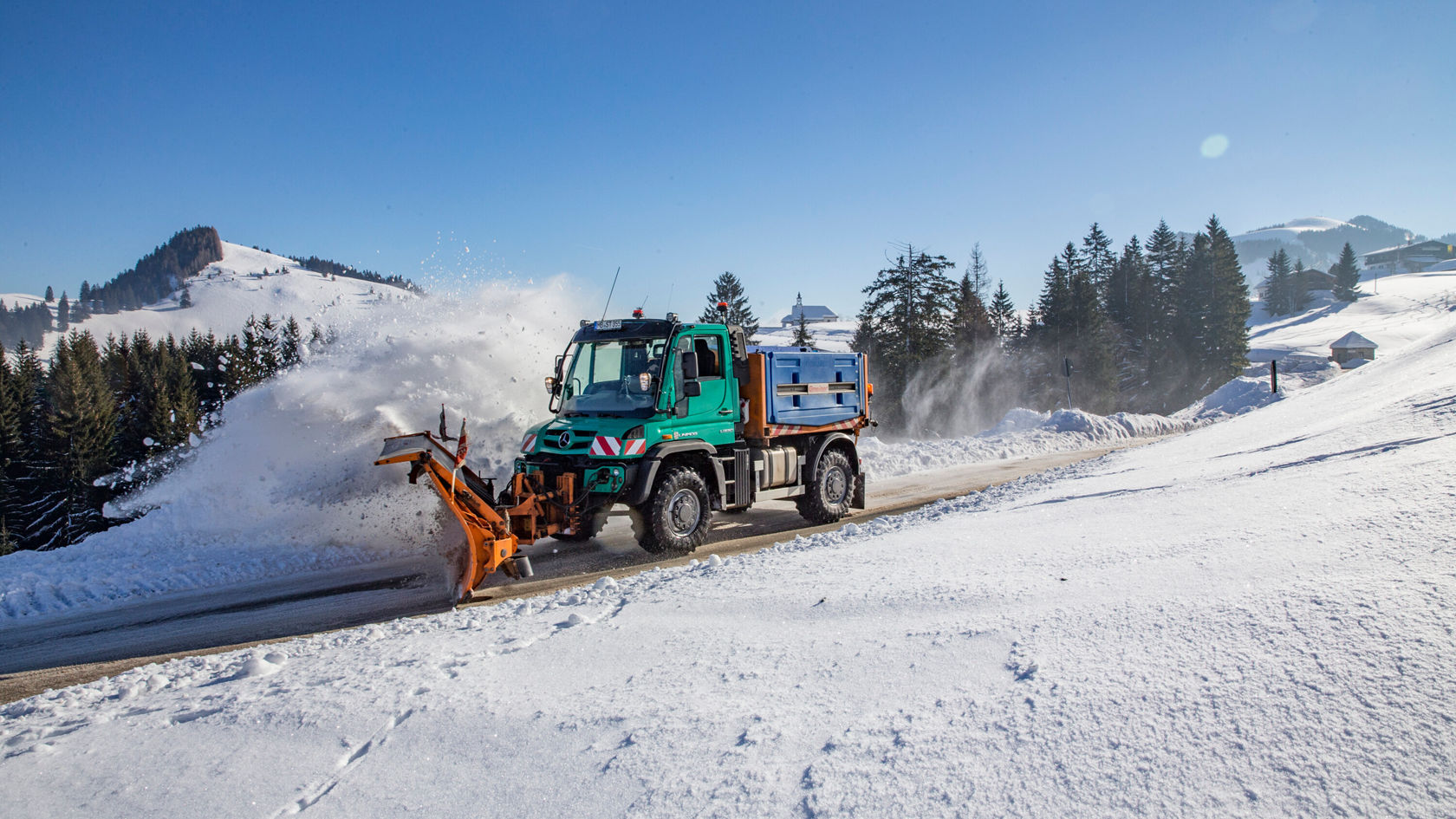 Un véhicule Mercedes-Benz équipé d'une lame de déneigement dégage une route enneigée dans un paysage montagneux, tandis que la neige est repoussée sur le côté.