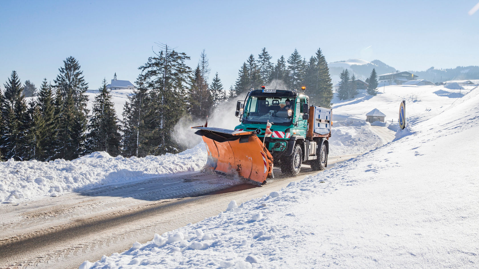 Un chasse-neige Mercedes-Benz dégage une route enneigée dans un paysage hivernal, entouré de sapins, avec des montagnes en arrière‑plan.
