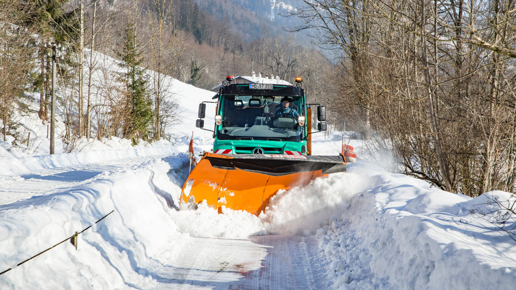 Un chasse-neige Mercedes-Benz dégage un chemin enneigé, tandis que des congères sont visibles de chaque côté. L'environnement présente un paysage hivernal avec des arbres.