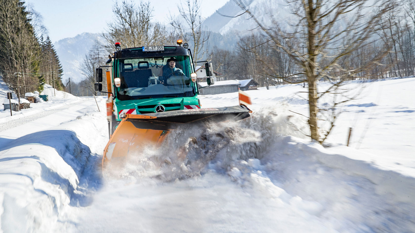 Un chasse-neige Mercedes-Benz dégage une route enneigée dans un paysage hivernal, avec des montagnes en arrière-plan. Le conducteur manœuvre activement le véhicule.