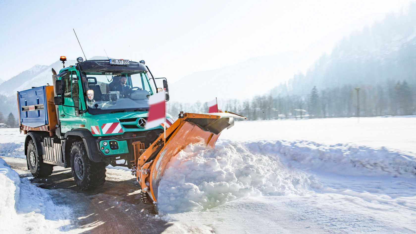 Un camion Mercedes-Benz équipé d'une lame de déneigement déblaye une route enneigée dans un environnement montagneux. Le camion est vert et orange, tandis que le paysage est marqué par la neige et les montagnes.