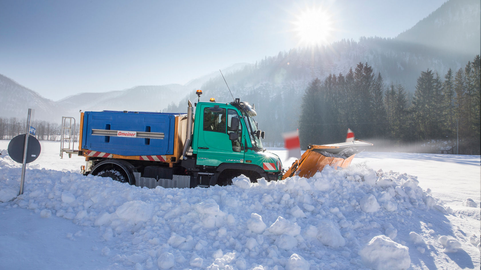 Un camion Mercedes-Benz équipé d'un chasse‑neige déblaye une étendue enneigée dans un paysage hivernal, avec des montagnes en arrière‑plan et un ciel ensoleillé.