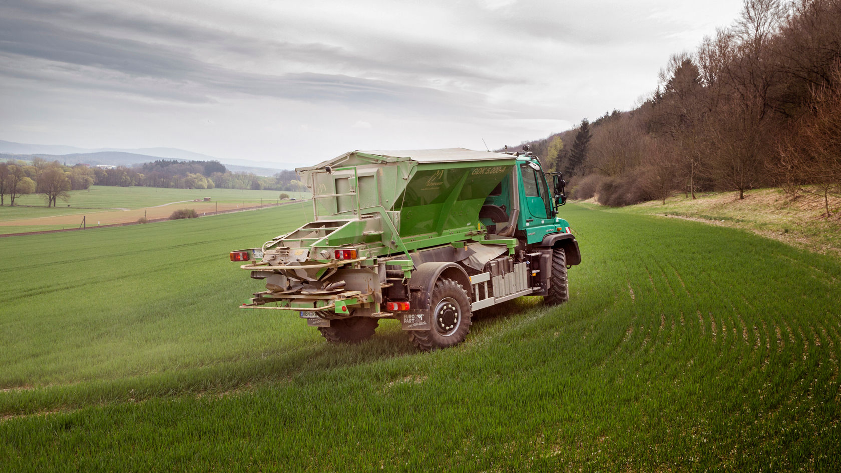 Un camión agrícola verde circula por un campo de trigo mientras distribuye fertilizante. En segundo plano se ven colinas suaves y una zona boscosa.