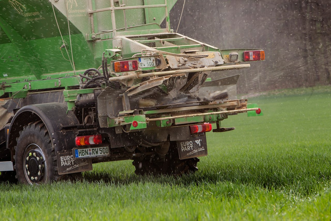 Un vehículo agrícola pulveriza líquido sobre un campo verde. La vista trasera muestra el equipamiento especial y la matrícula del vehículo, que se emplea para las labores de campo.