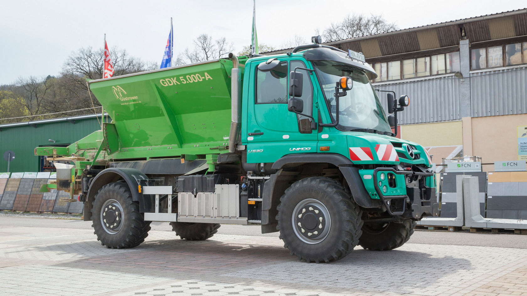Un Unimog Mercedes-Benz verde con una gran plataforma de carga está sobre una superficie pavimentada, listo para trabajos de transporte o de intervención en la construcción o la agricultura.