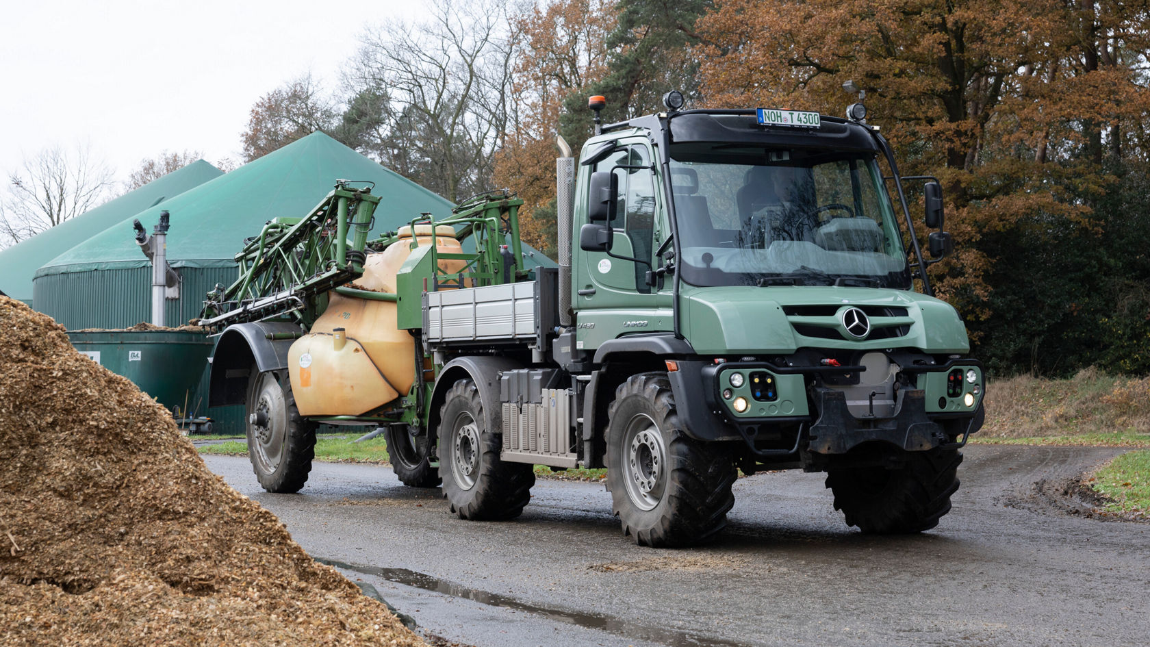 A green Unimog with a field sprayer drives on a country road. Warehouses can be seen in the background.