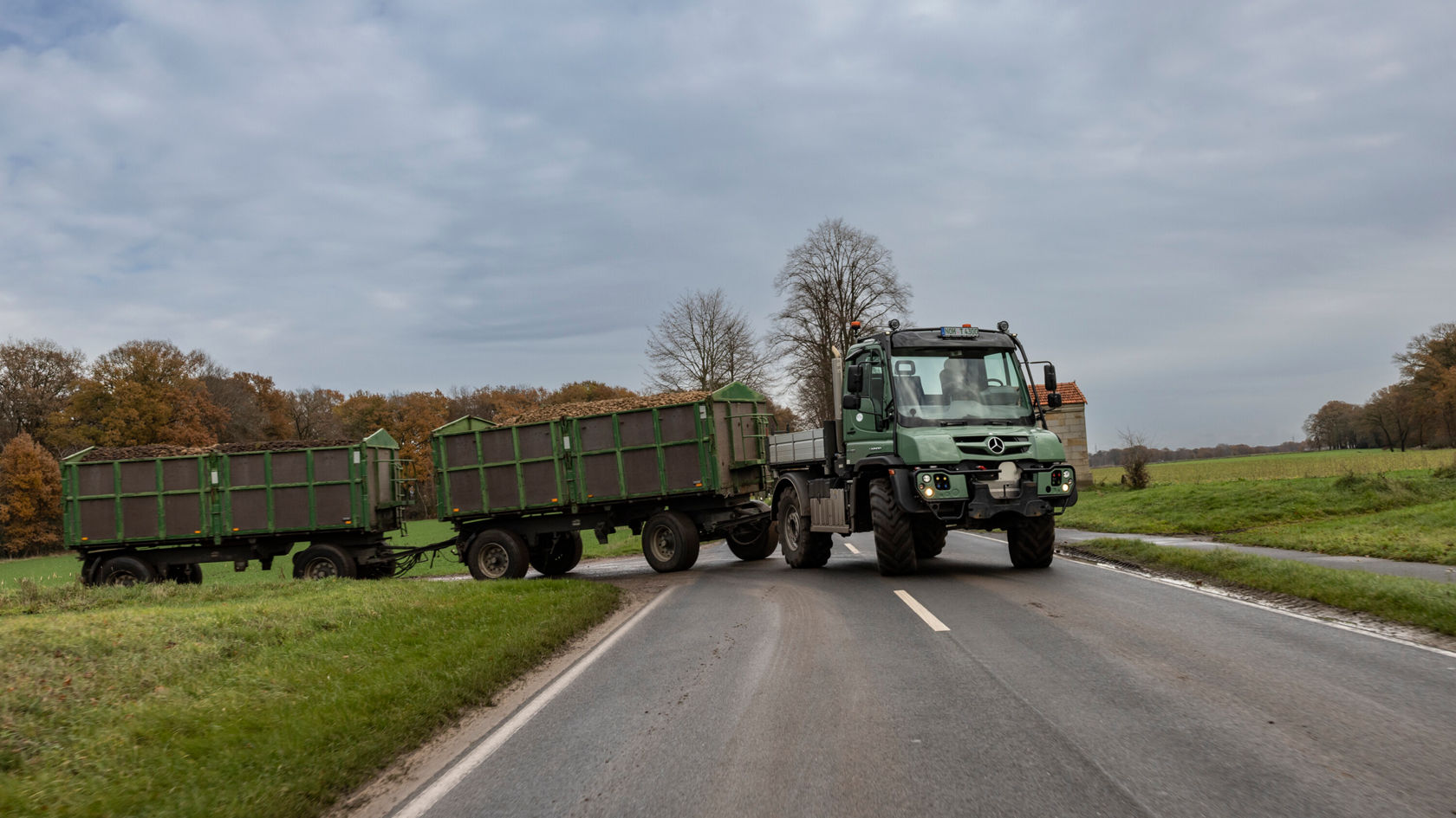 A green Unimog with two trailers stands at a road junction. The trailers are loaded with wood or agricultural materials.