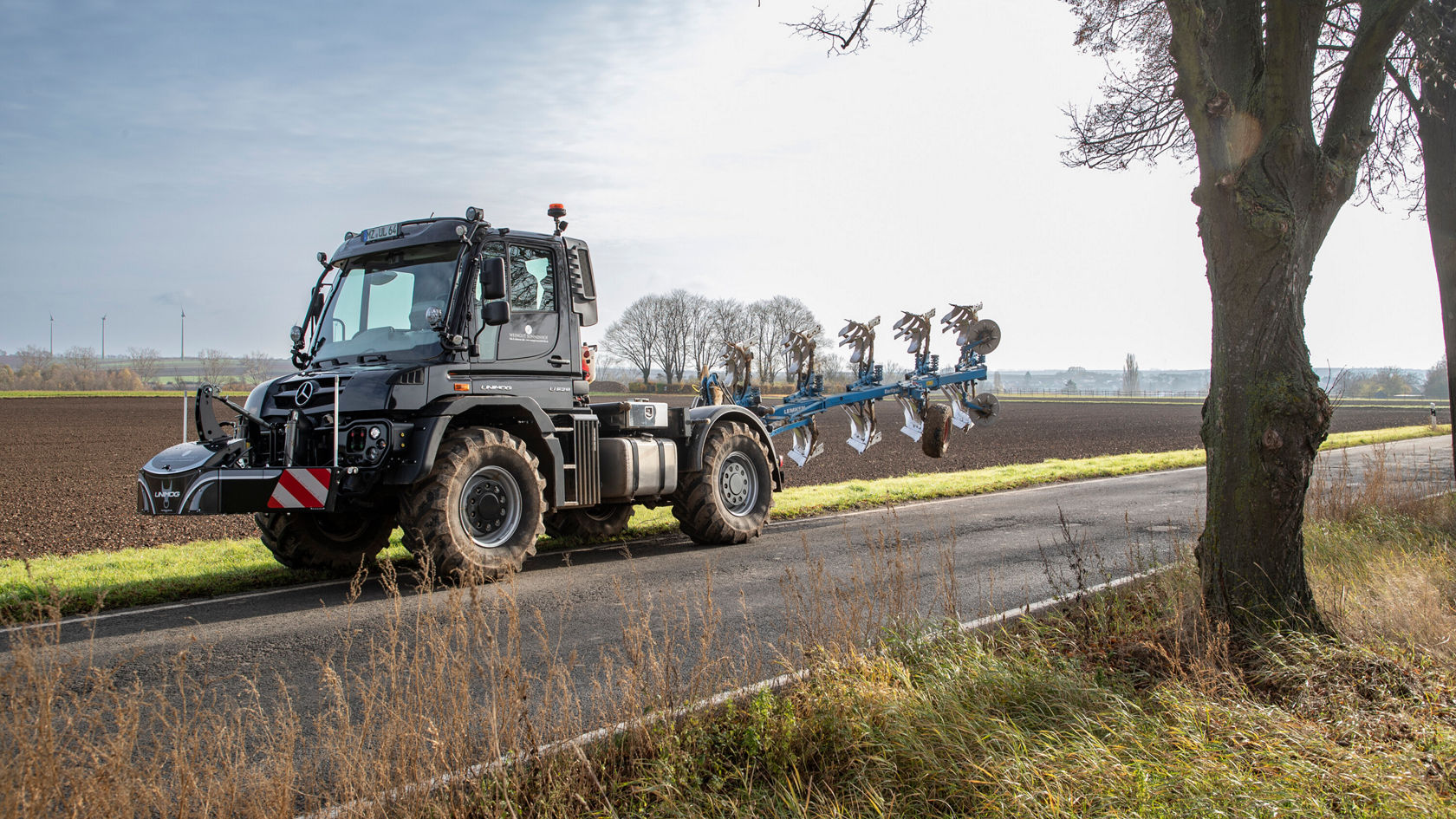 Un tracteur Mercedes‑Benz roule sur une route de campagne et traîne une charrue équipée de plusieurs socs. En arrière-plan, des champs agricoles sont visibles.