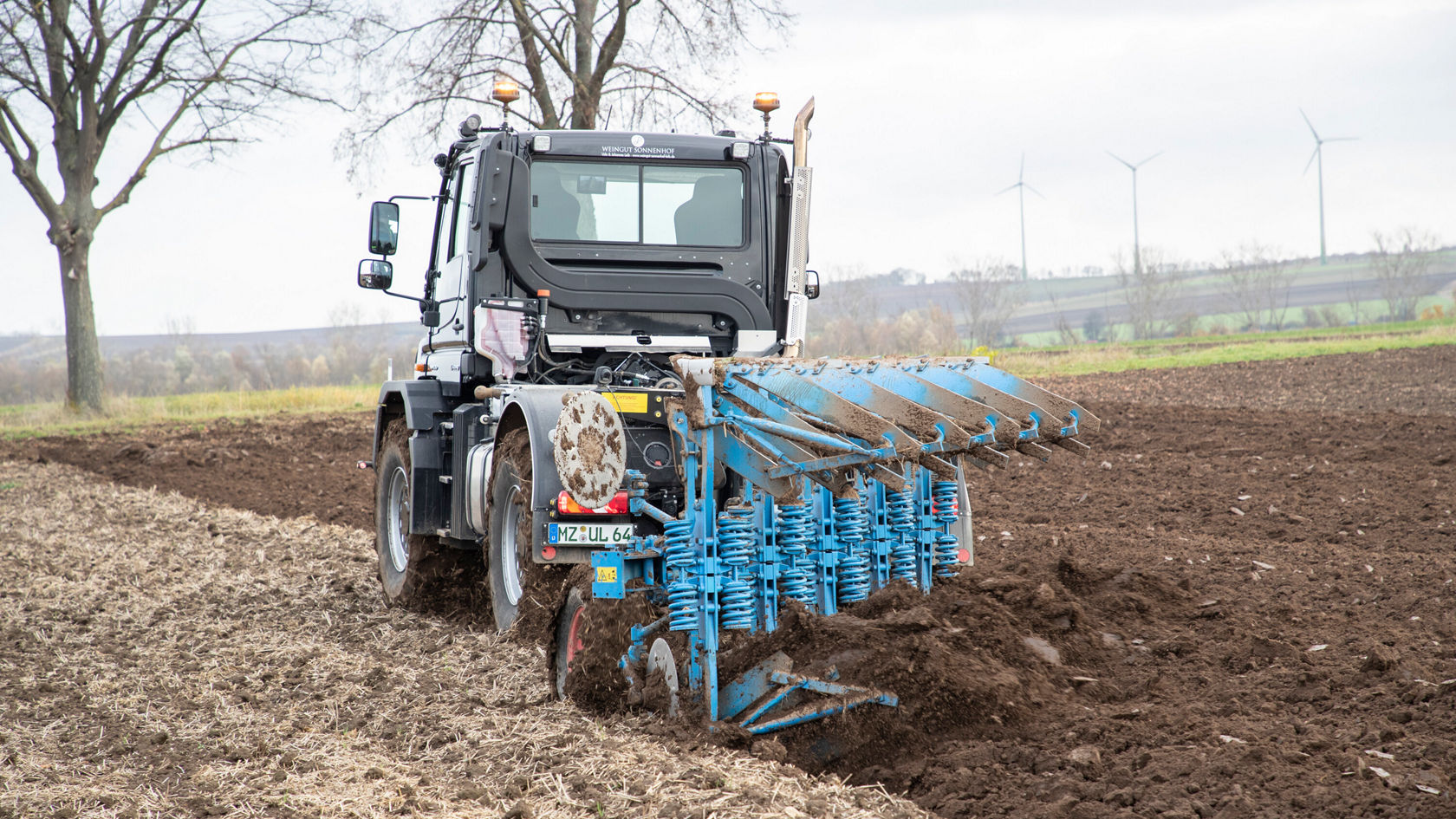 Un tracteur agricole Mercedes‑Benz, équipé d'une charrue bleue, travaille un champ fraîchement labouré. Des éoliennes sont visibles à l'arrière-plan.