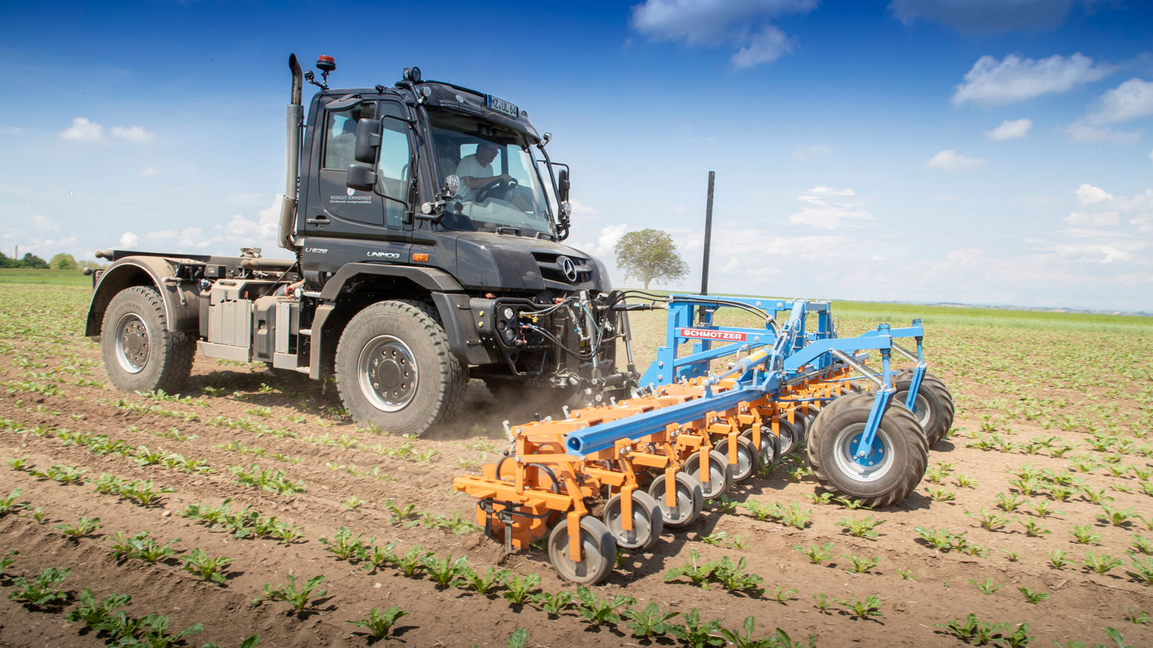 Un camion Mercedes‑Benz travaille un champ à l'aide d'un équipement agricole. La machine laboure le sol dans ce champ verdoyant sous un ciel dégagé.