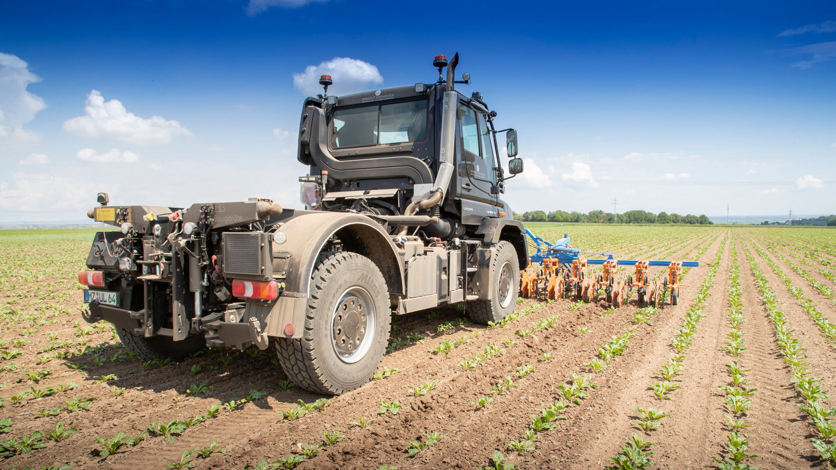 Un tracteur Mercedes‑Benz travaille un champ de plantes vertes. Le tracteur est vu de dos et tire une charrue.