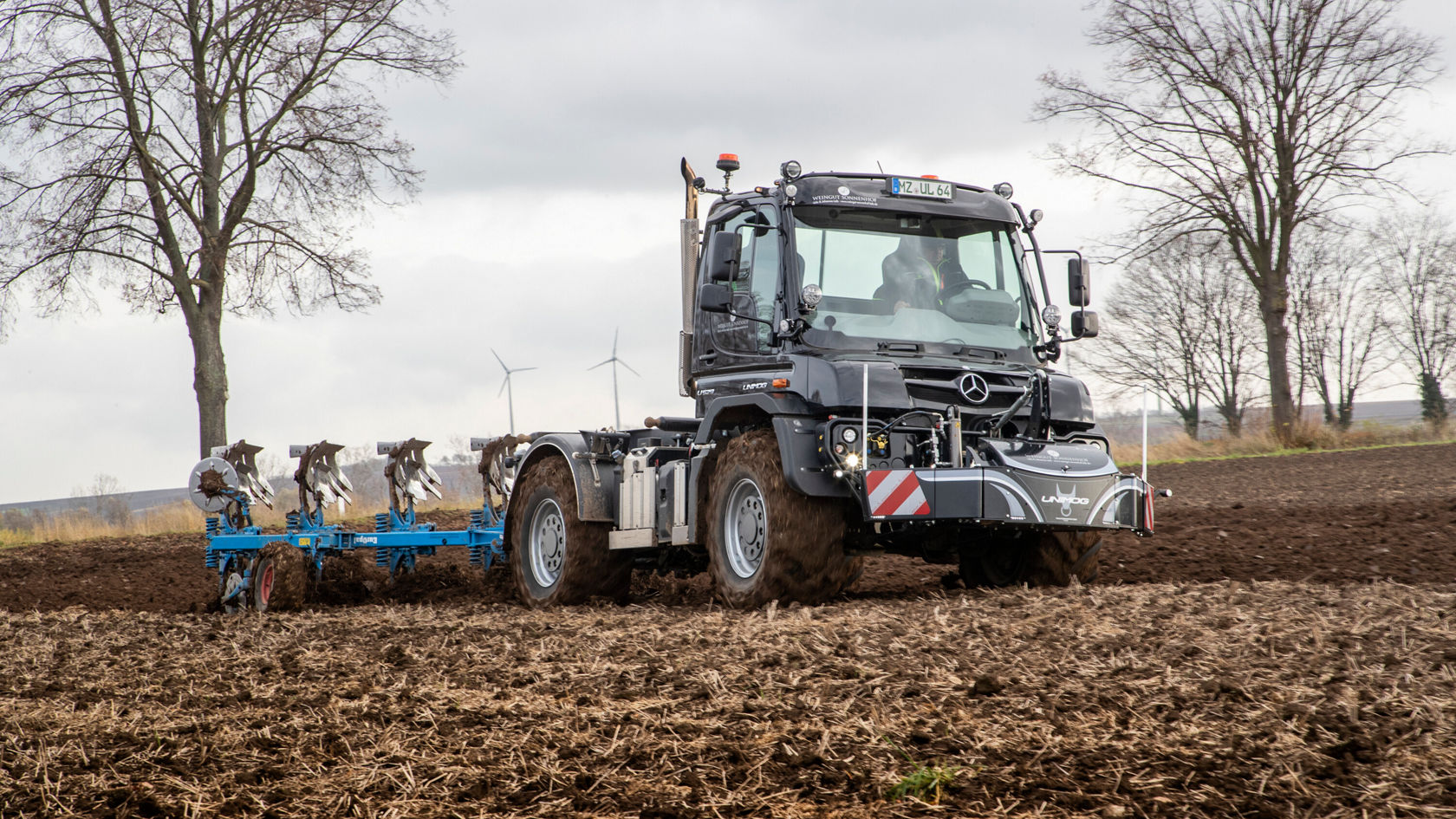 Un tracteur Mercedes‑Benz laboure un champ avec une charrue. À l'arrière‑plan, on voit des arbres et des éoliennes.