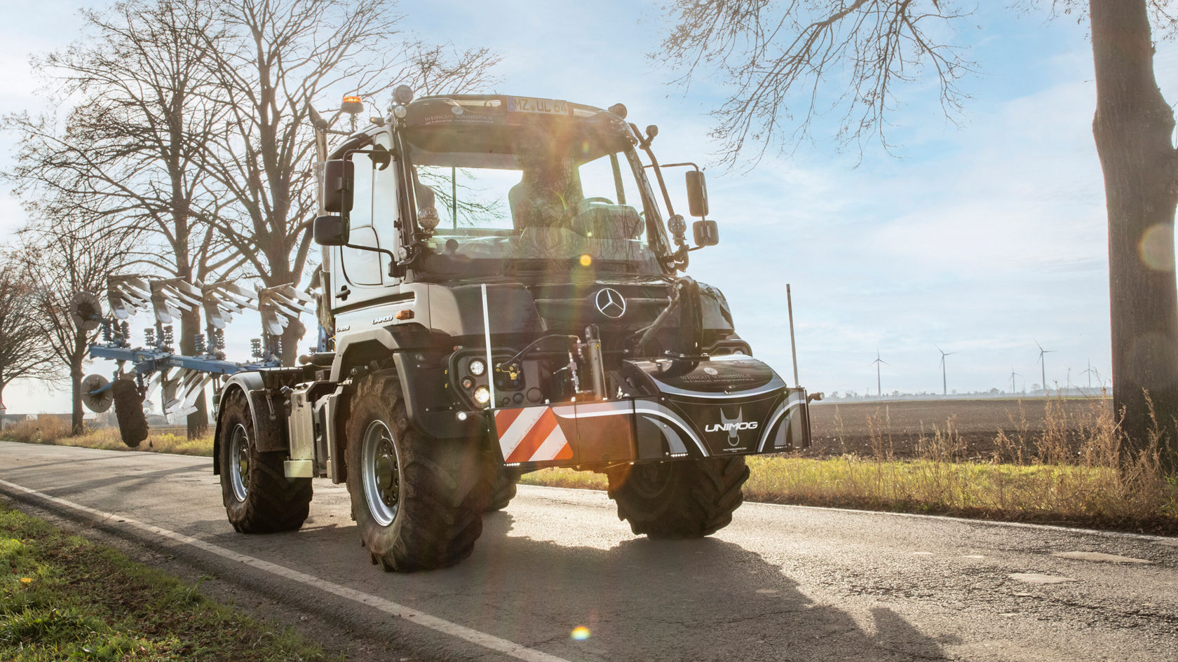 Un tracteur Mercedes‑Benz roule sur une route de campagne, entouré d'arbres, équipé d'outils agricoles à l'arrière, idéal pour des travaux agricoles.