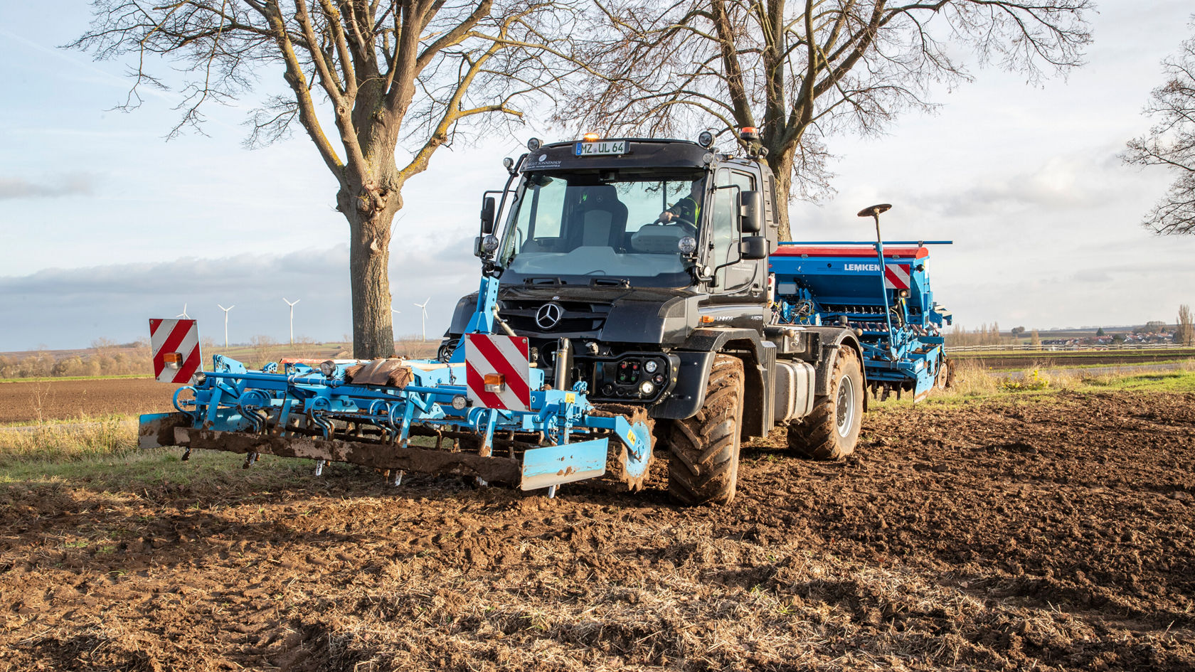 Un tracteur Mercedes‑Benz noir travaille un champ avec une machine agricole bleue, tandis que des arbres et des éoliennes sont visibles en arrière-plan.