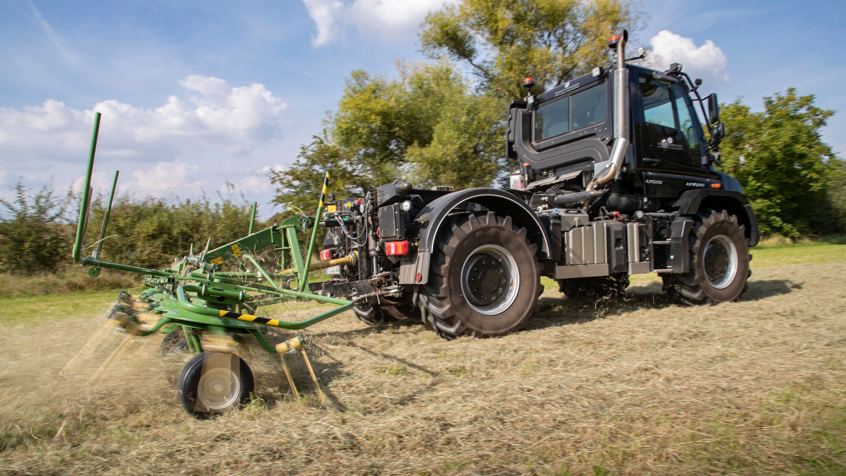 Un tracteur Mercedes‑Benz travaille un champ avec un outil de retournement vert. La machine présente des pneus robustes et des équipements techniques modernes par temps ensoleillé.
