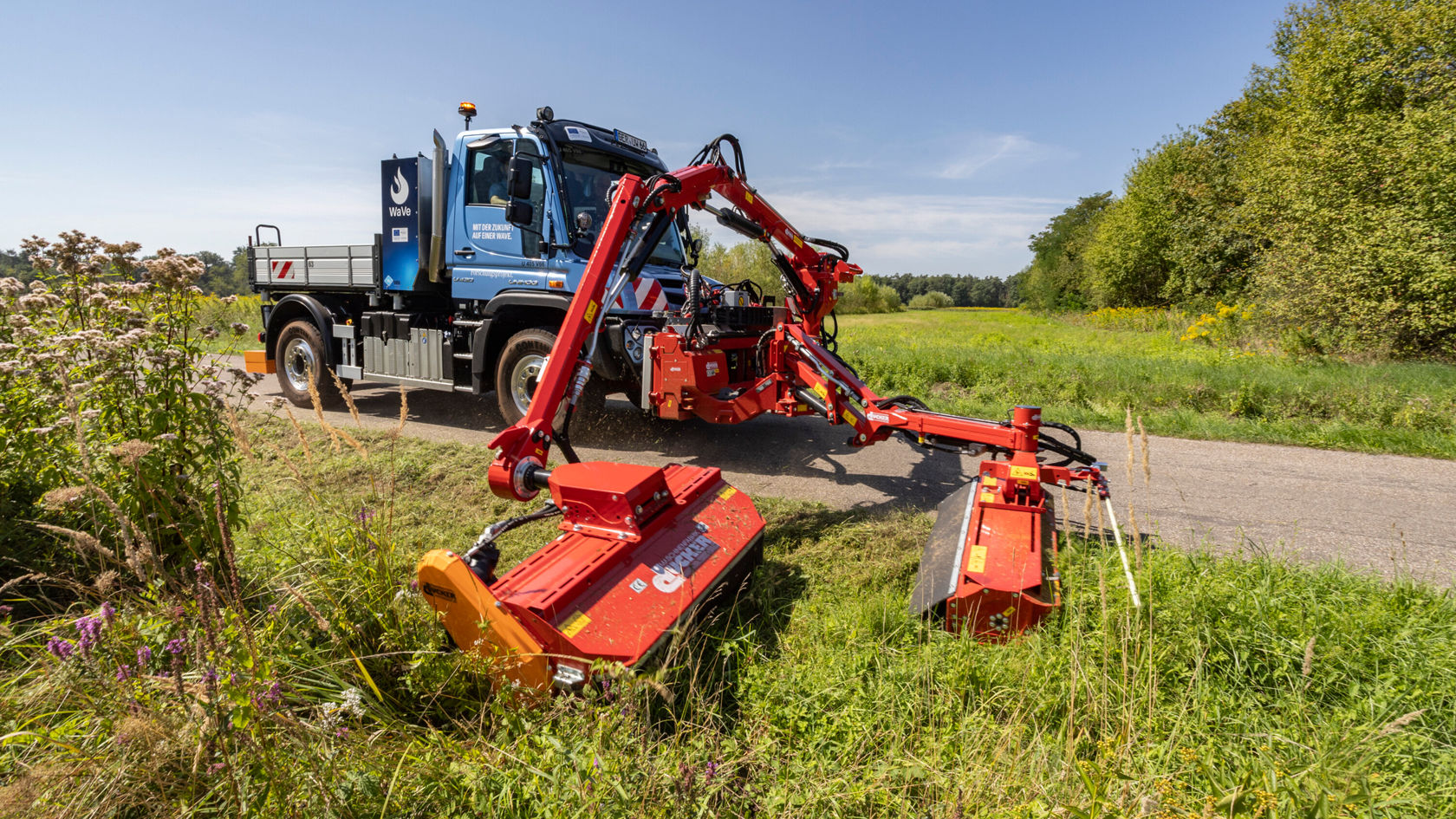 Ein Mercedes-Benz Unimog mit einer rotierenden Mähmaschine auf der Straße, umgeben von Grünflächen und Bäumen, während der Fahrer die Maschine bedient.