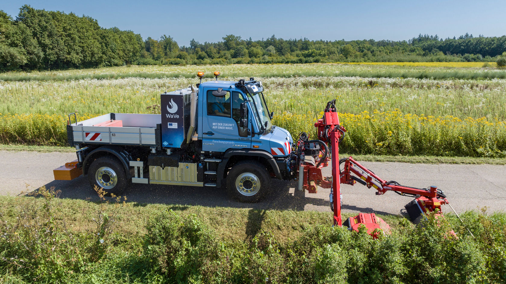 Ein Mercedes-Benz Unimog steht an einer Landstraße mit einem roten Arbeitsarm, der zur Pflege und Wartung von Vegetation eingesetzt wird.