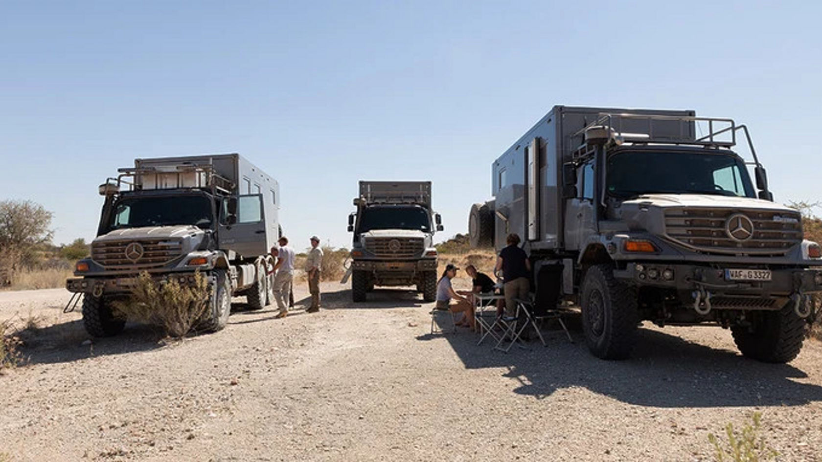 Drei Mercedes-Benz Zetros stehen auf einer Sandstraße. Dazwischen sitzen Personen auf Klappstühlen.