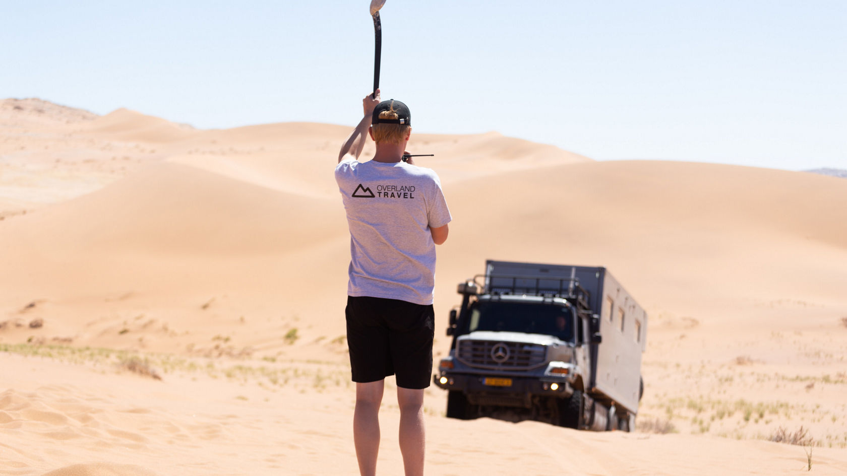 Eine Person steht in einer Wüstenlandschaft, während ein Mercedes-Benz Zetros durch den Sand fährt.