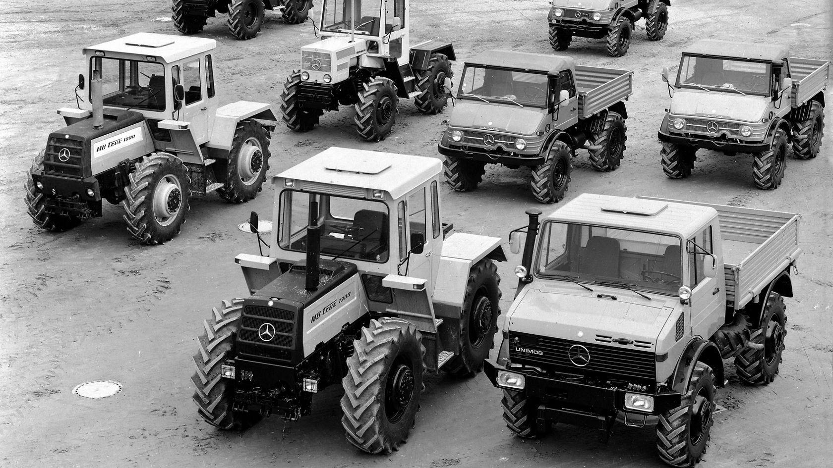 Mehrere Unimog-Fahrzeuge von Mercedes-Benz stehen auf einem Platz. Die Fahrzeuge sind in schwarz-weiß und zeigen verschiedene Modelle, die für vielfältige Anwendungen ausgelegt sind.
