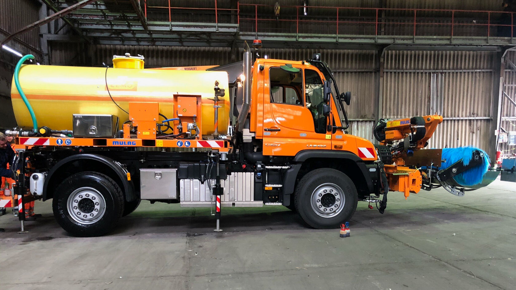 Een oranje Mercedes-Benz Unimog, uitgerust met een grote gele tank en een zijarm, staat in een industriële hal.
