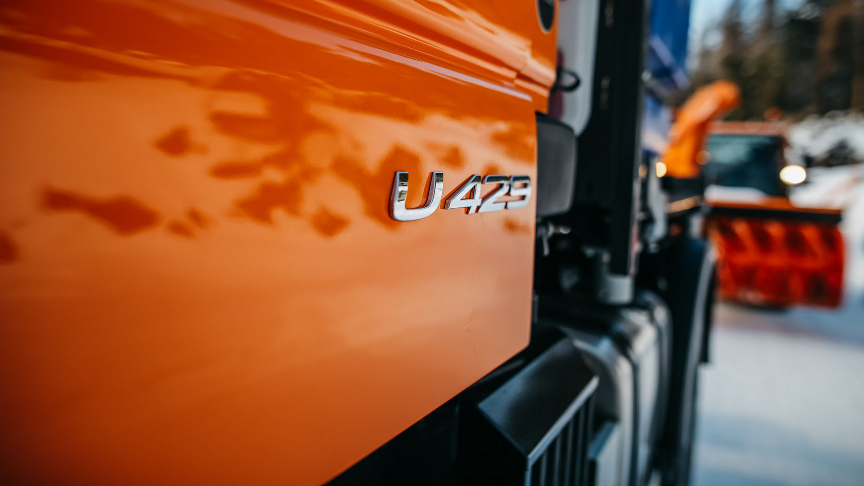 U423 model designation on the side panel of an orange Mercedes-Benz Unimog; a white snowplough is visible in the background.