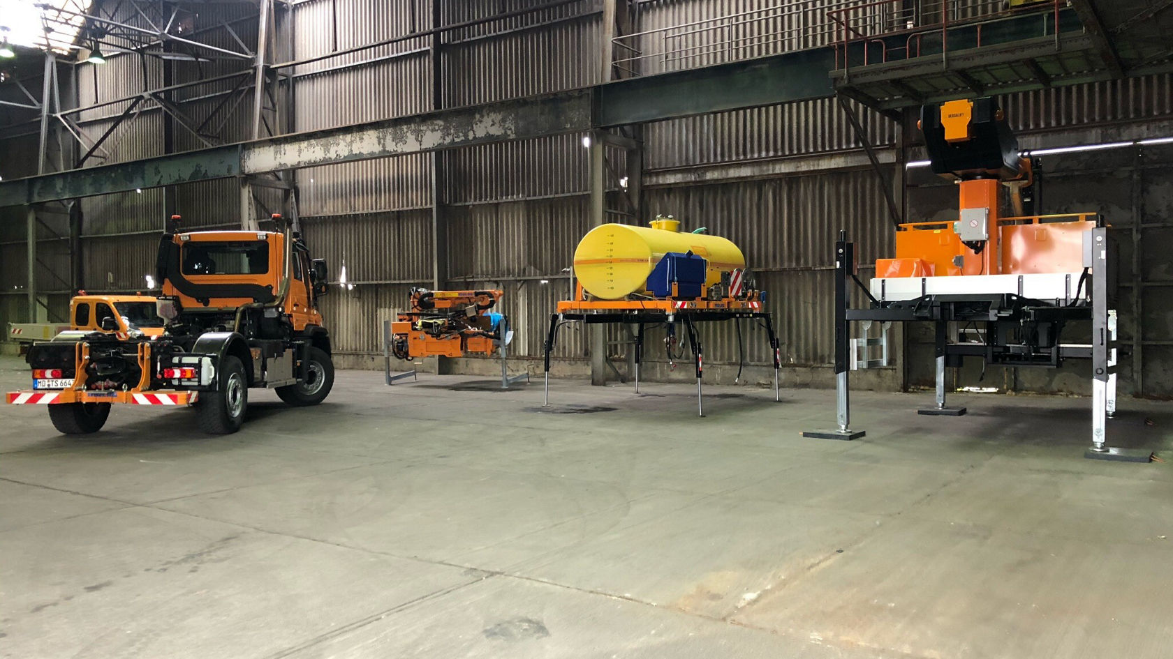 Een Mercedes-Benz Unimog staat in een loods naast andere apparaten, waaronder een gele watertank en een oranje machine.