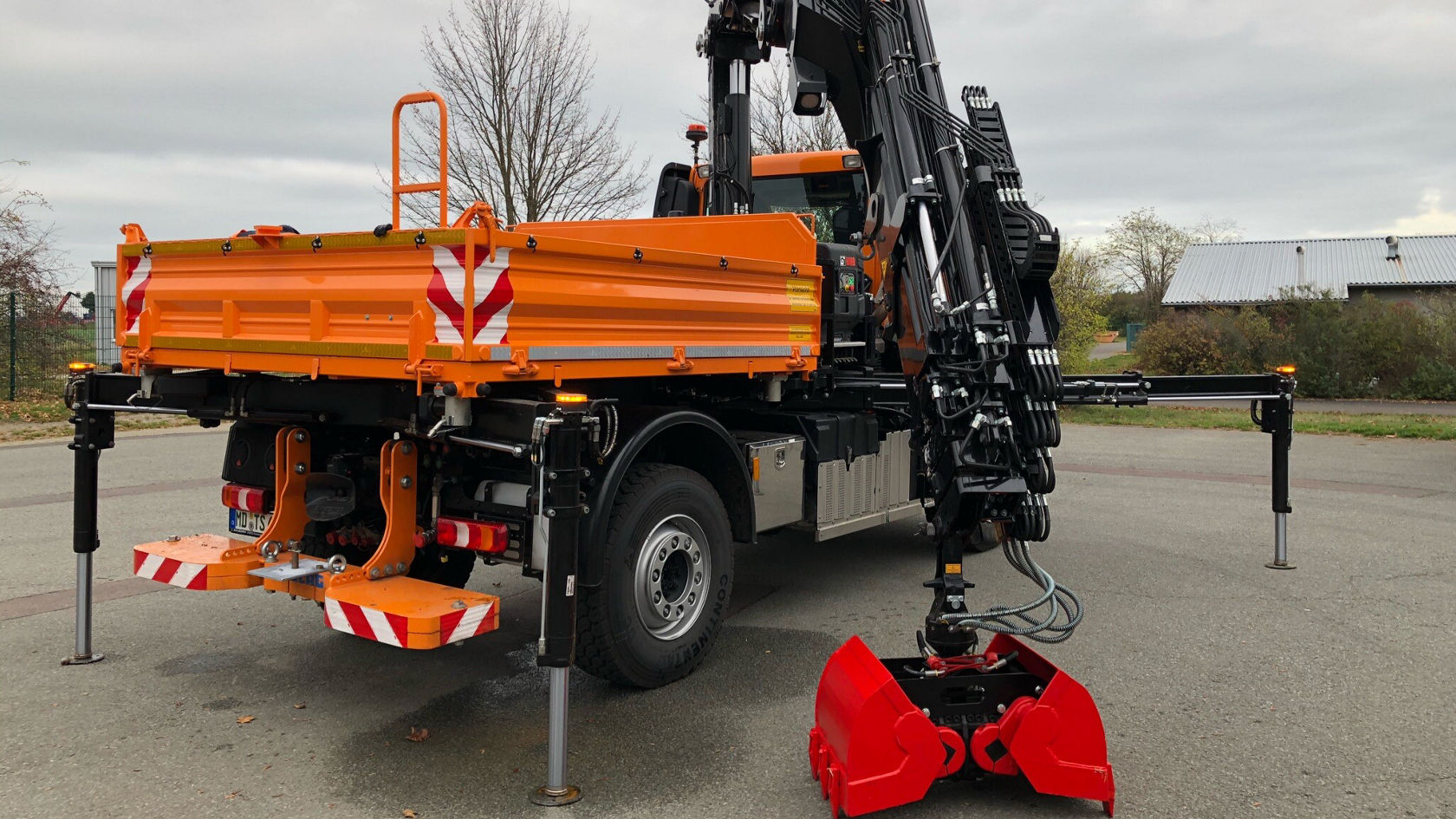 Een Mercedes-Benz Unimog met een oranje opbouw en kranen, uitgerust met een rode grijper, staat op een parkeerplaats.