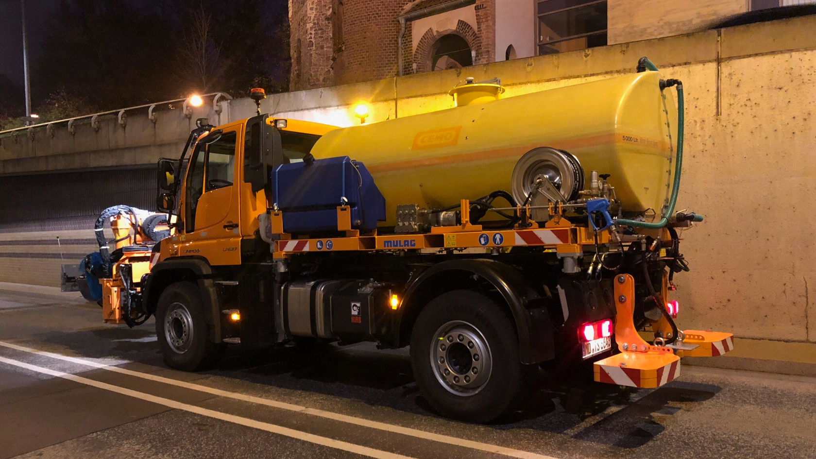 Een Mercedes-Benz Unimog staat langs een weg, uitgerust met een gele tank en verschillende gereedschappen op de laadbak, klaar om 's nachts wegonderhoudswerkzaamheden uit te voeren.