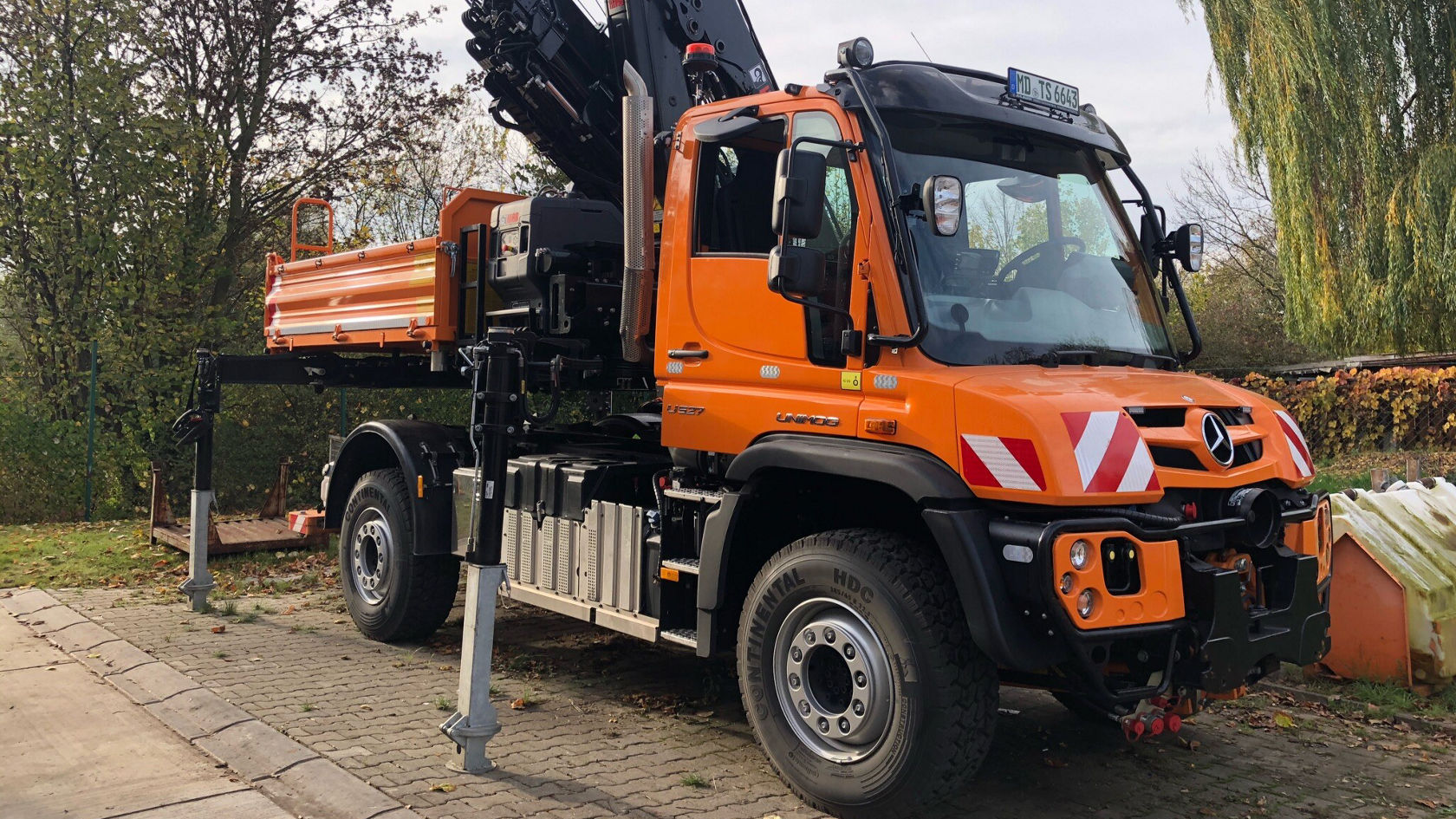 Een oranje Mercedes-Benz Unimog met een kraanopbouw staat op een verharde plaats. De kraan is uitgeklapt en klaar voor inzet.