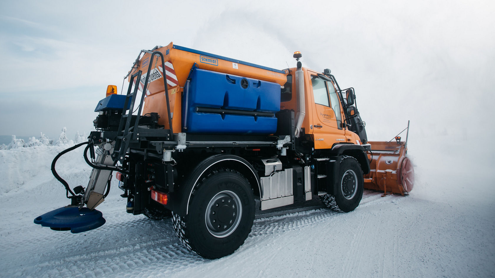 A Mercedes-Benz Unimog clears snow with a snowplough on a snow-covered road. The orange truck is shown from the rear with a spreader attachment.