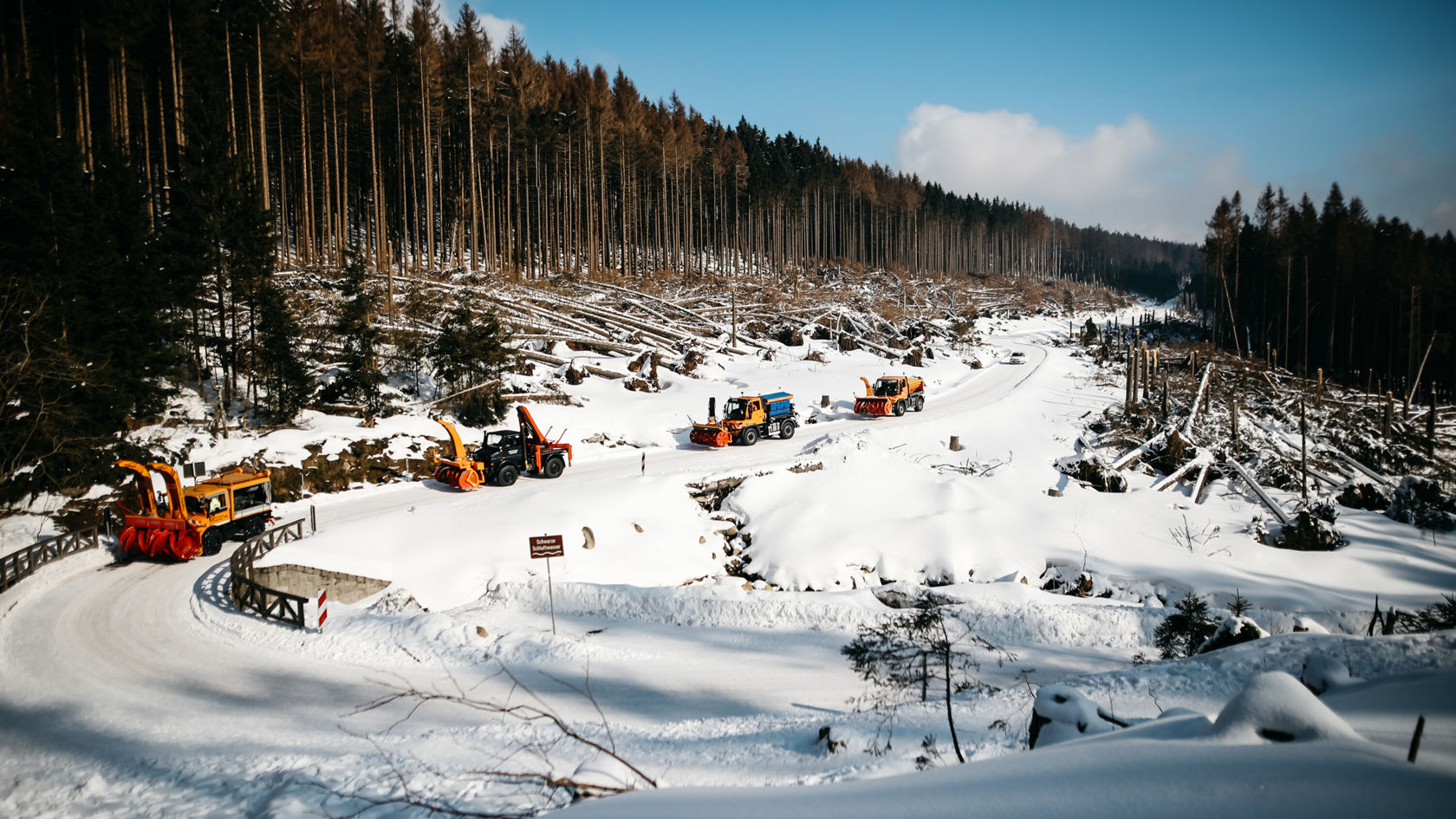 Snow-clearing vehicles, including a Unimog from Mercedes-Benz, are working on a snow-covered road in a forest area with felled trees in the background.