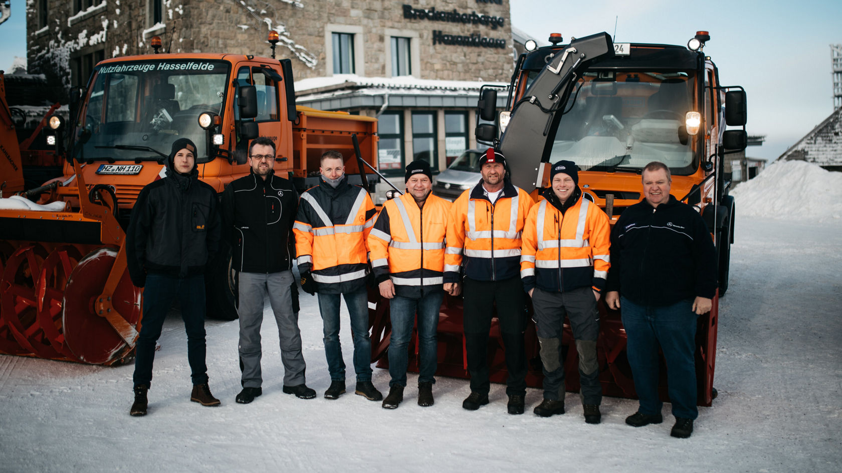 A group of six people in orange work jackets next to two orange Mercedes-Benz Unimog snow-clearing vehicles in the snow.