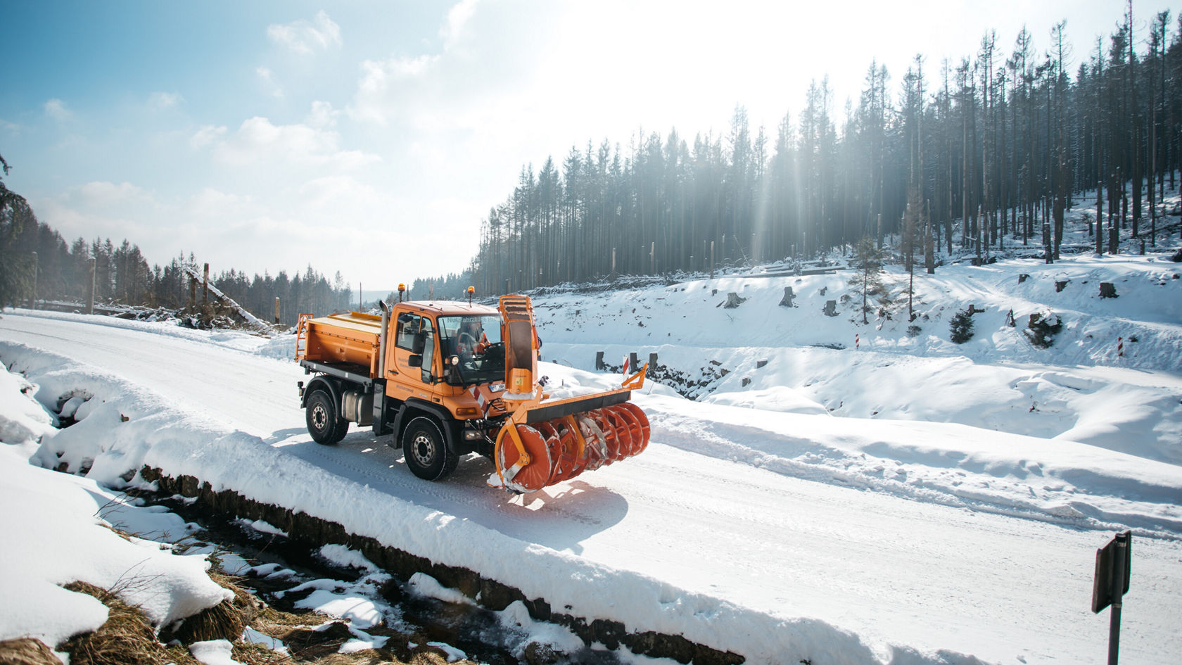 A Mercedes-Benz Unimog clears a snow-covered road in a forested area with a snowplough. The surroundings are wintry, with snow and trees in the background.