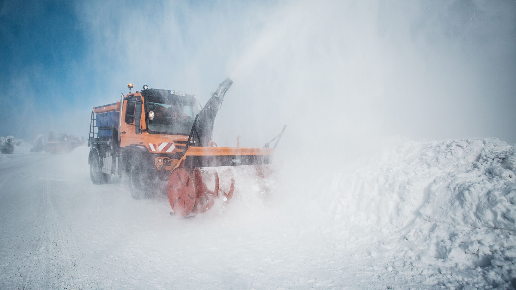 An orange Mercedes-Benz Unimog with a snowplough clears snow from a snow-covered road as snow is kicked up in front of the vehicle.