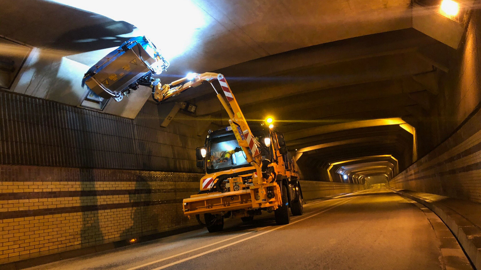 Een Mercedes-Benz Unimog staat in een tunnel en heft met een uitschuifbare arm een container omhoog. De tunnel is zwak verlicht en toont een geasfalteerde weg.