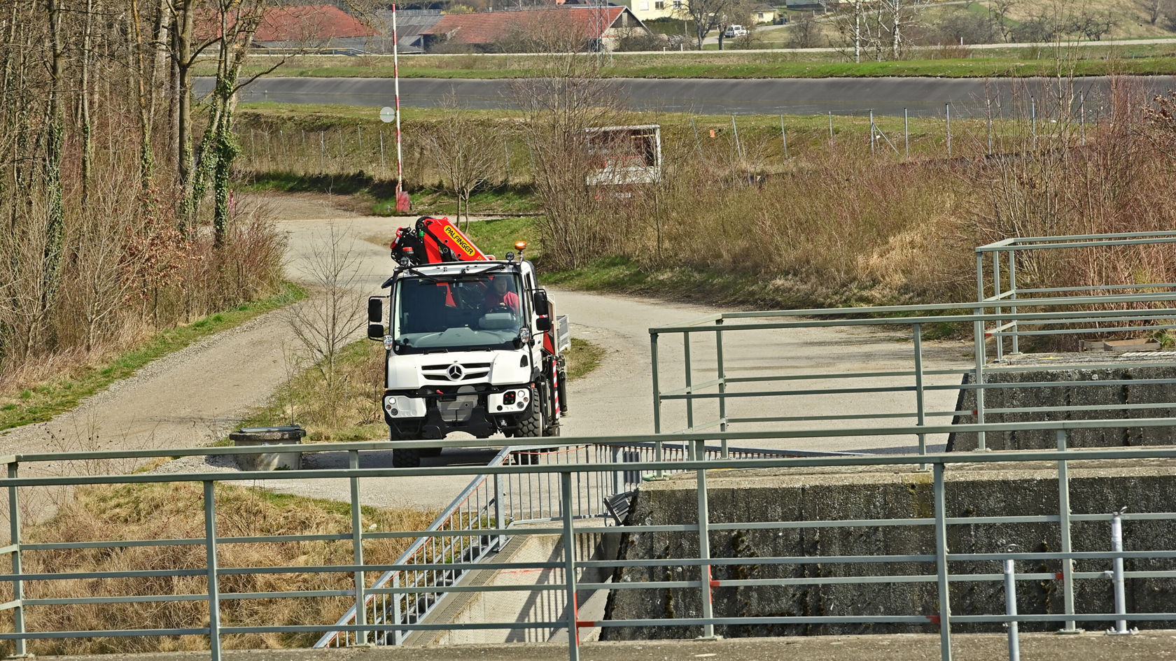 Ein Mercedes-Benz Unimog fährt auf einem schmalen Weg, während er mit einer Ladung ausgestattet ist. Im Hintergrund sind Zäune und abgesperrte Bereiche sichtbar.