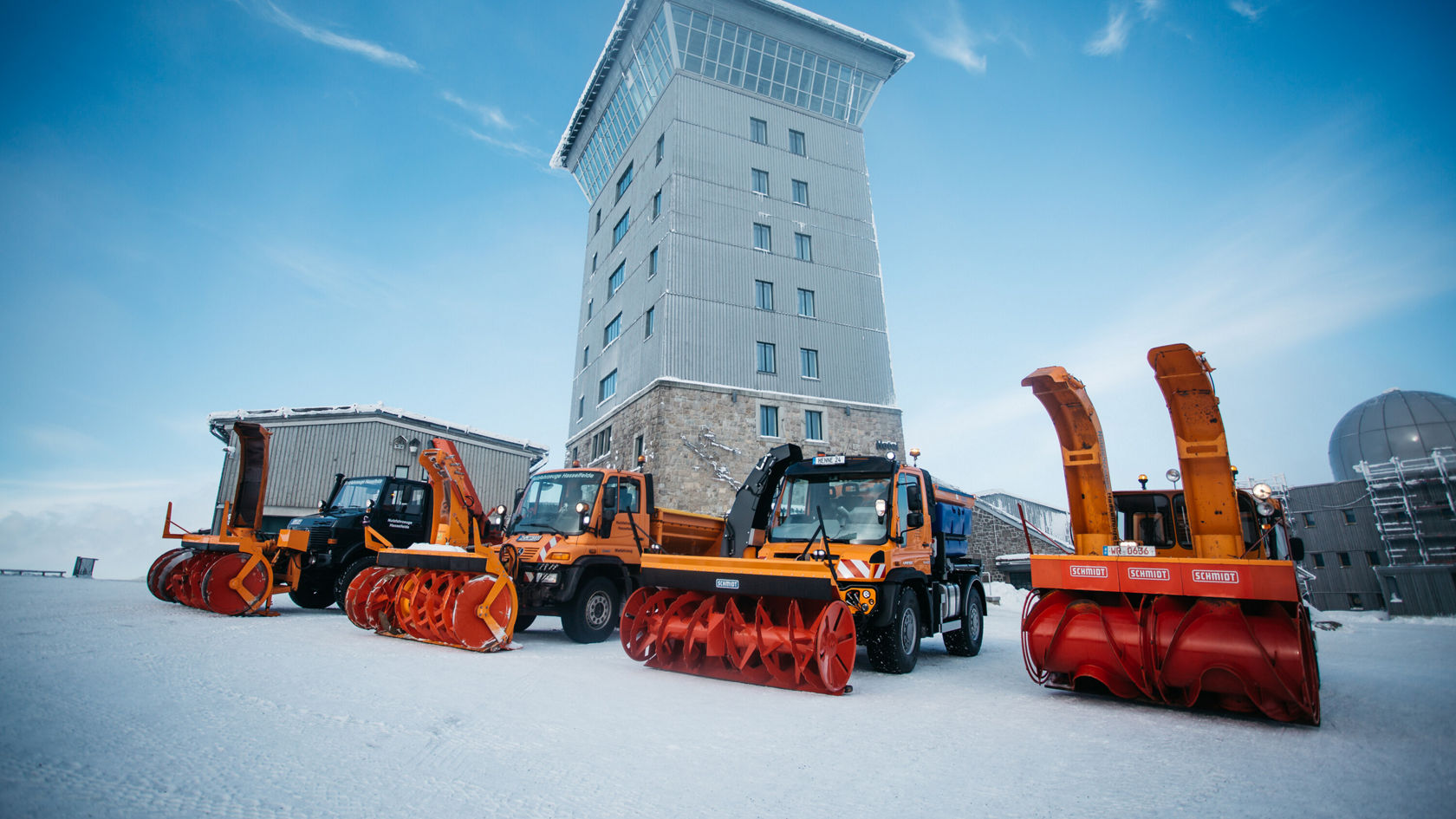 A Mercedes-Benz Unimog with a snowplough stands in the foreground, next to two other emergency vehicles at the foot of a tower in a snow-covered environment.