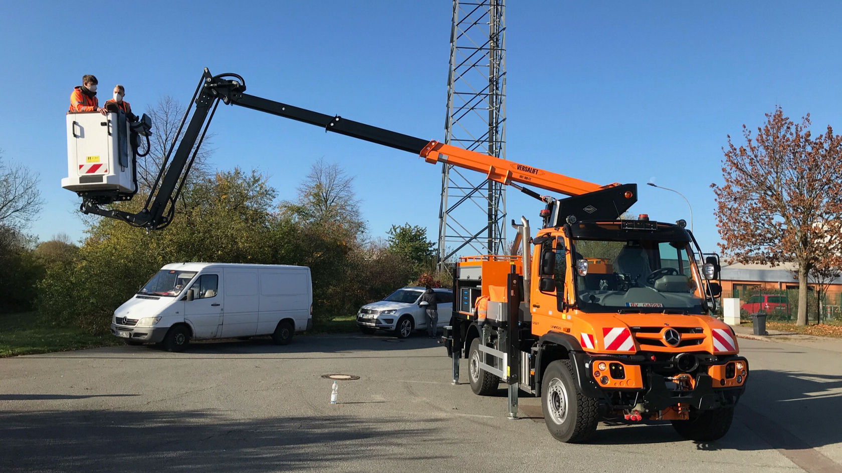 Een Mercedes‑Benz Unimog met een knikarm die een werkplatform optilt, waarop twee personen zitten, staat naast een witte bestelwagen en een ander voertuig.