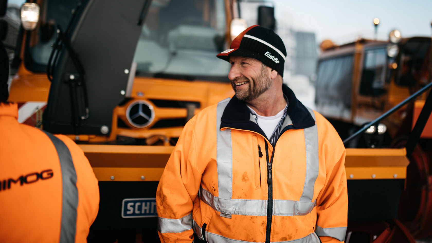 A Mercedes-Benz Unimog truck is in the background, while a man in reflective work clothing and a cap smiles and talks with other people.