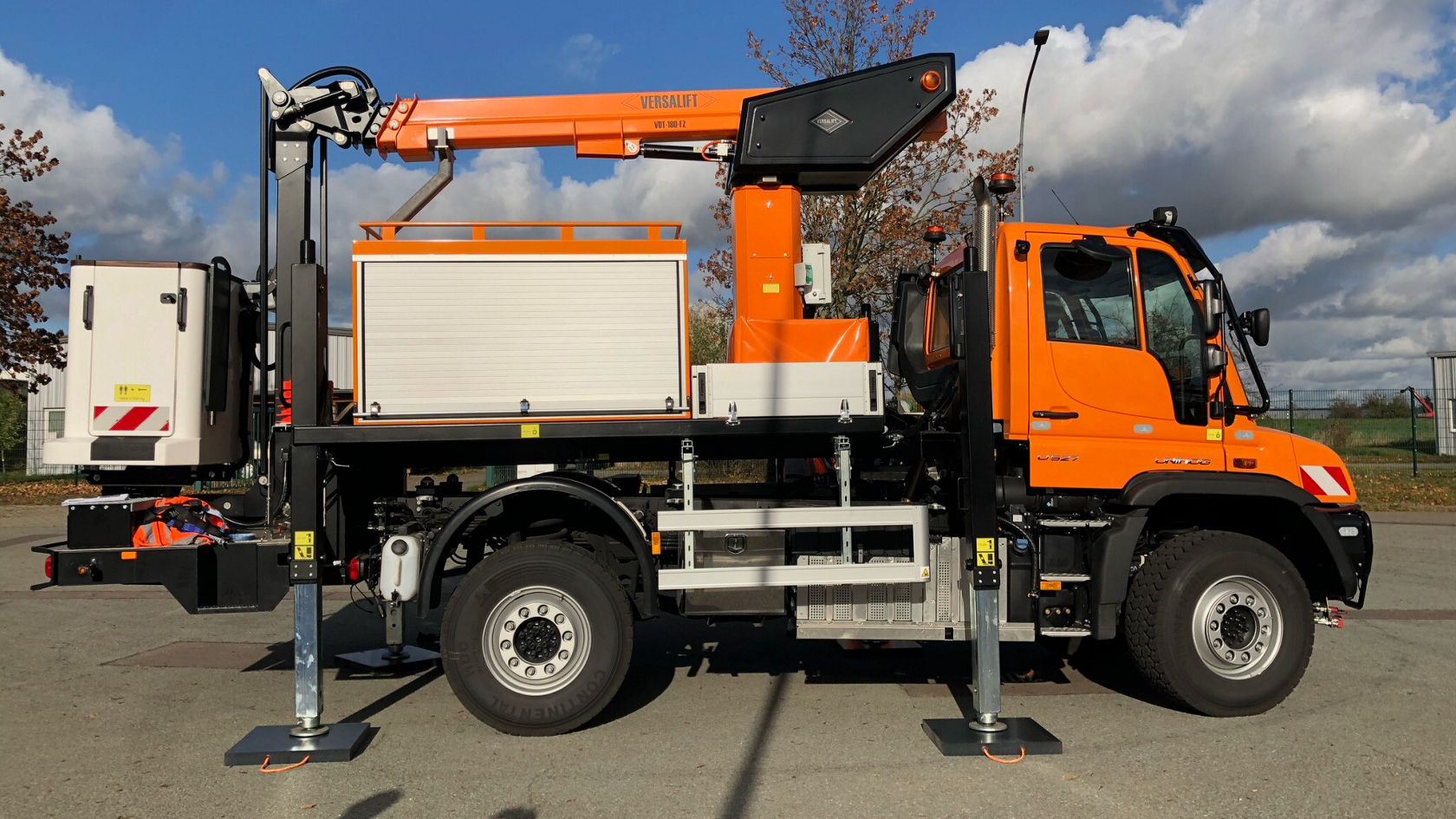 Een Mercedes-Benz Unimog in fel oranje met een werkbak en een kraanarm, gestabiliseerd door steunpoten, staat op een geplaveide ondergrond.