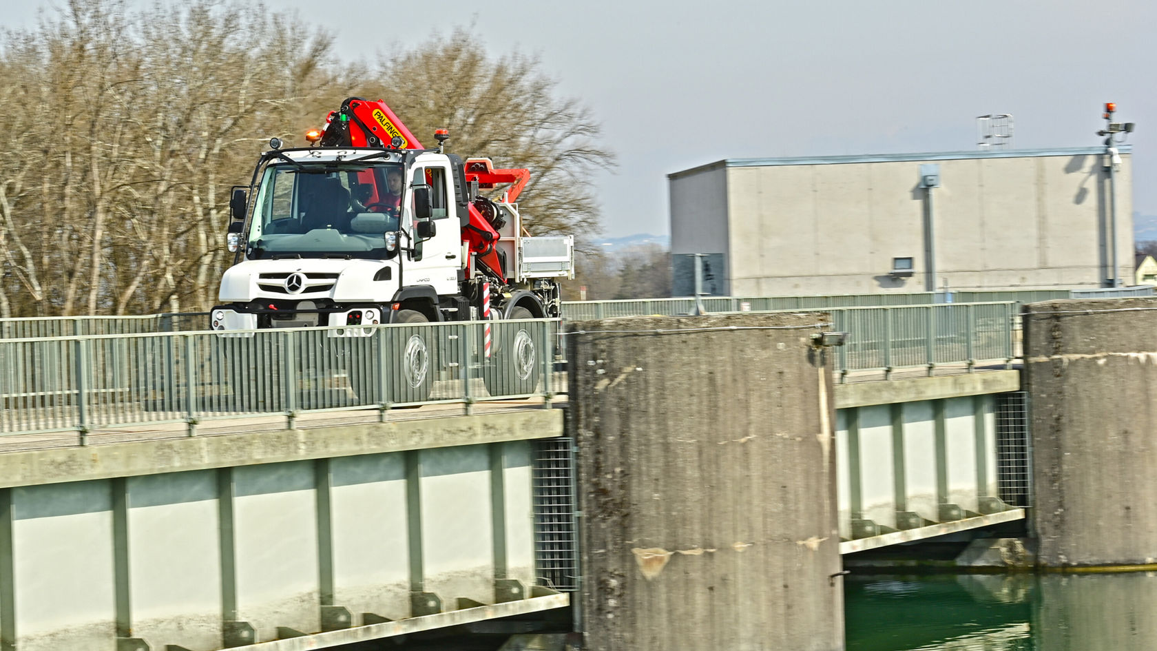 Ein Mercedes-Benz Unimog fährt auf einer Brücke über eine Wasseroberfläche, mit einer roten Ladekran-Ausrüstung auf der Ladefläche.