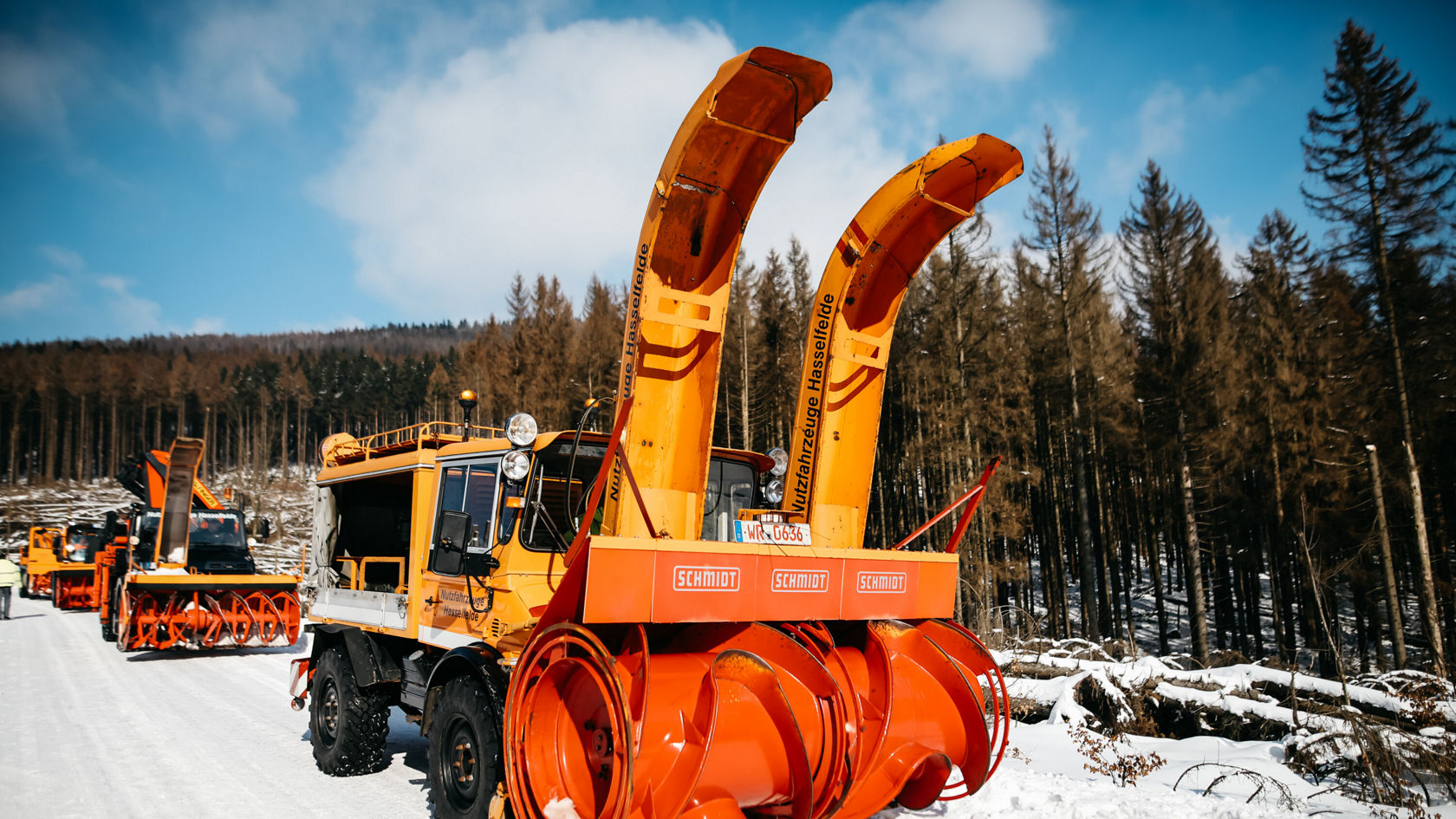 A Mercedes-Benz Unimog with two large orange snow blowers stands on a snow-covered path, surrounded by bare trees in the background.
