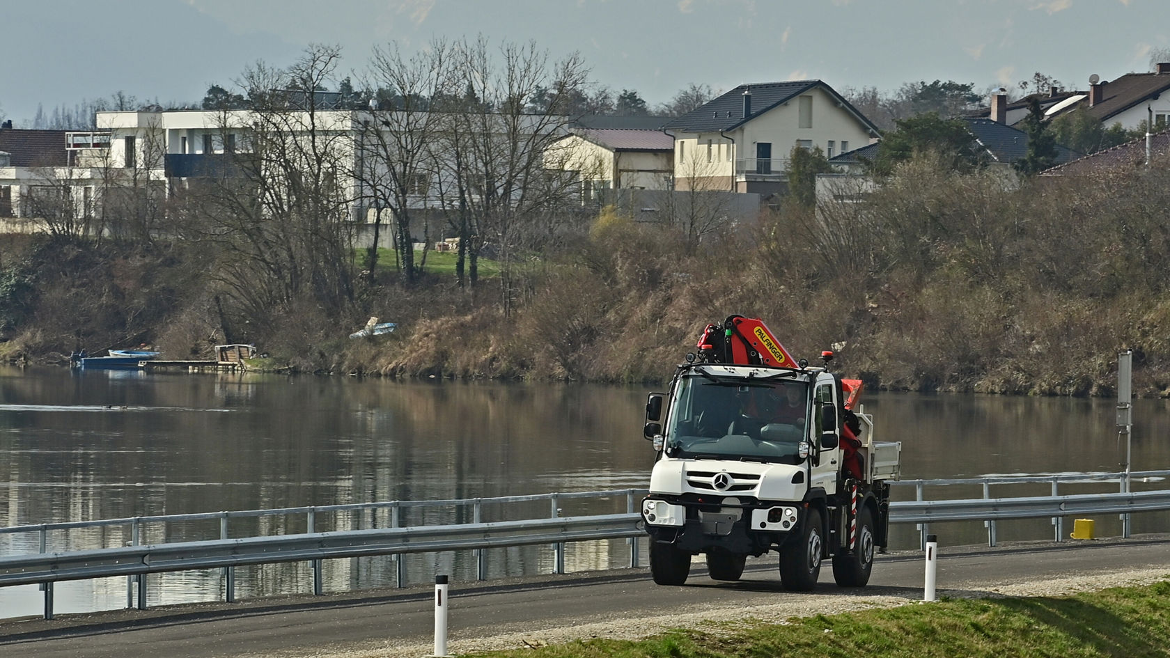 Ein Mercedes-Benz Unimog fährt auf einer Straße entlang eines Gewässers, während im Hintergrund Wohnhäuser und Bäume sichtbar sind.